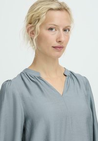 Hellgraue, leichte Bluse mit V-Ausschnitt und gerafftem Ärmel-Design. Glatte Textur mit fließendem Schnitt.