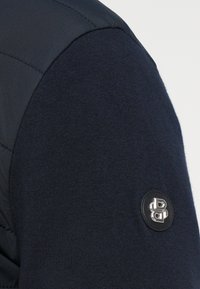 Tumši zils piedurknēm nesaturošs džemperis ar teksturētu vilnas adījuma korpusu un gludu, izšūtu plecu daļu, rotāts ar melnu ādas aplīti ar iegravētu logotipu.