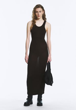 Junge Frau mit langen lockigen Haaren, die ein schwarzes ärmelloses Maxikleid, schwarze Stiefel trägt und eine schwarze Handtasche hält.