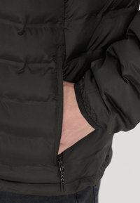 Giacca nera isolata con tasca con zip, polsini elasticizzati e una texture liscia e opaca. Primo piano sulla zona del lato e della manica.