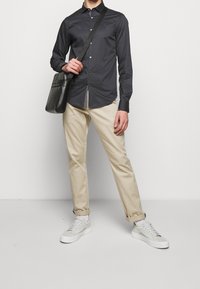 Mörkgrå långärmad skjorta med knappar, beige byxor, vita och ljusgrå sneakers, bär en svart axelväska. Enkelt mönster och passform.