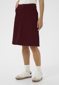 Jupe en velours côtelé bordeaux au design droit, avec poches avant et fermeture bouton. Portée avec des baskets blanches et des chaussettes côtelées.