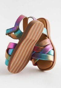 Sandales taille enfant avec semelles marron et brides métalliques colorées en nuances de rose, violet, bleu et or, posées sur un fond blanc.