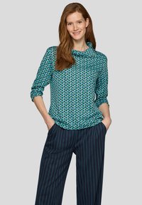 Frau mit langen roten Haaren, die ein blaues Oberteil mit geometrischem Muster und dunkelblaue, weit geschnittene Nadelstreifenhosen trägt und mit den Händen in den Taschen lächelt.