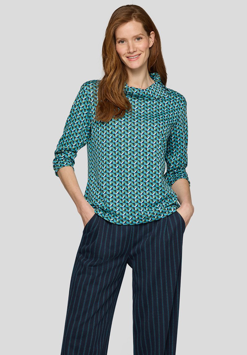 Frau mit langen roten Haaren, die ein blaues Oberteil mit geometrischem Muster und dunkelblaue, weit geschnittene Nadelstreifenhosen trägt und mit den Händen in den Taschen lächelt.