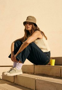 Cappello a secchiello beige chiaro, top senza maniche crema testurizzato, jeans svasati blu scuro, sneakers verdi con suola rialzata e tazza gialla sui gradini.