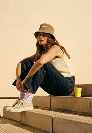 Un chapeau seau beige clair, un haut sans manches crème texturé, un jean évasé bleu foncé, des baskets vertes avec une semelle surélevée, et une tasse jaune sur des marches.