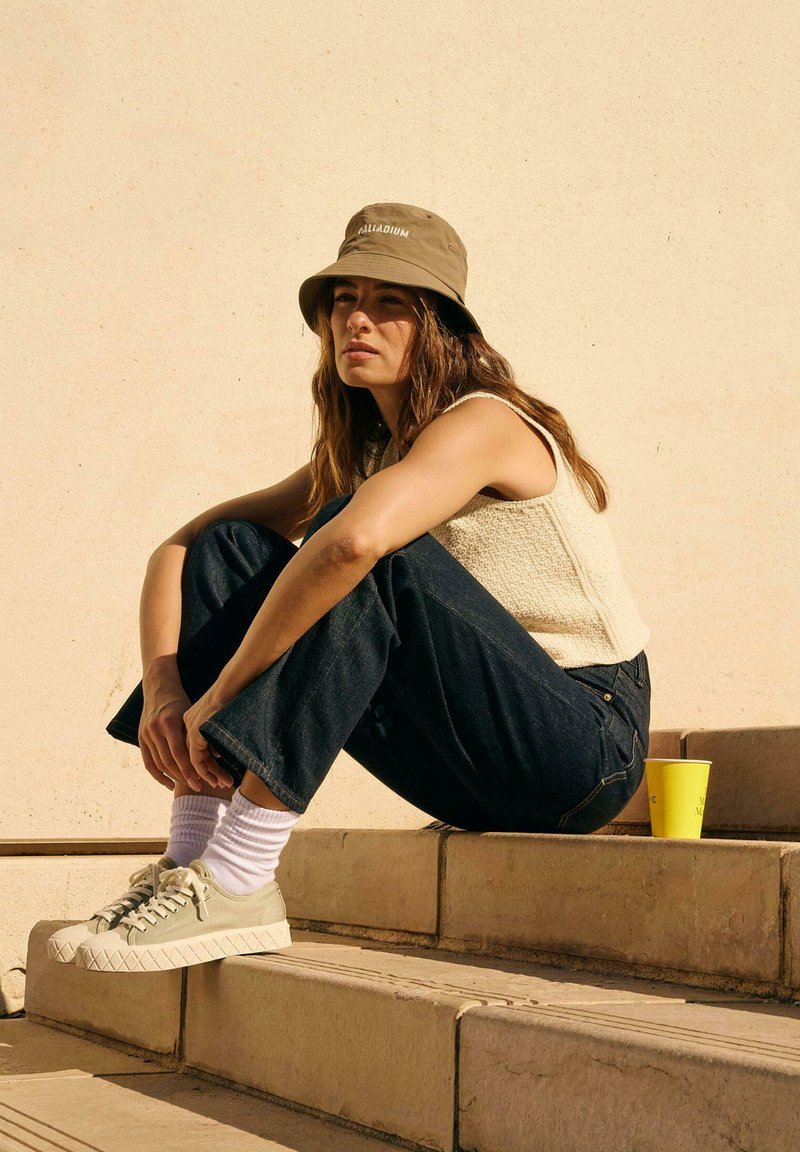 Cappello a secchiello beige chiaro, top senza maniche crema testurizzato, jeans svasati blu scuro, sneakers verdi con suola rialzata e tazza gialla sui gradini.