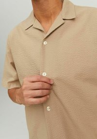 Camicia beige a maniche corte con texture, con colletto da campo, parzialmente abbottonata, indossata da una persona che sta sistemando il primo bottone.