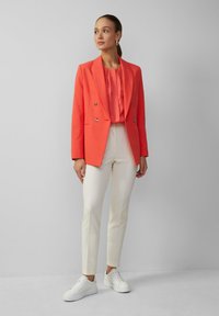 Korallefarbener doppelt geknöpfter Blazer, plissierte hellrosa Bluse, maßgeschneiderte cremefarbene Hose und weiße Sneaker. Glattes Material mit strukturierter Passform.