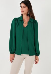 Blusa verde de manga larga con cuello, hombros abullonados y corte recto. Llevada sobre una camiseta de escote en V, combinada con pantalones de color claro.