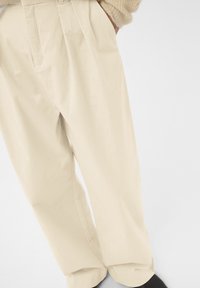 Pantaloni de culoare crem deschis, din bumbac, cu o croială lejeră, având pliuri frontale și buzunare laterale. Textură netedă și picior conic la capăt.