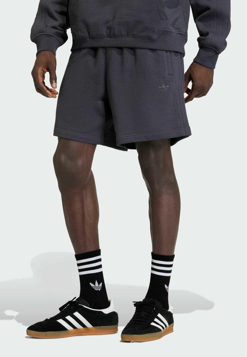 Donkergrijze katoenen shorts met een elastische tailleband, gecombineerd met zwarte Adidas schoenen met witte strepen en bijpassende zwarte sokken met strepen.