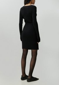 Schwarzes geripptes Kleid mit langen Ärmeln und taillierter Silhouette, kombiniert mit schwarzen Rüschenslippers und transparenten schwarzen Strumpfhosen.