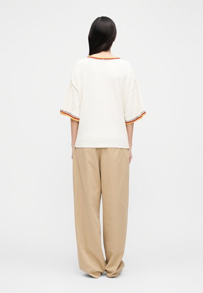 Biele nadrozmerné tričko s oranžovým lemom na výstrihu a rukávoch, kombinované s ľahkými béžovými širokými nohavicami. Jednofarebné pozadie.