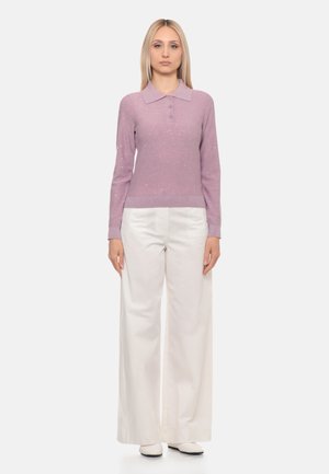Donna con capelli biondi lisci che indossa un maglione leggero viola chiaro a maniche lunghe e pantaloni bianchi a gamba larga, in piedi su uno sfondo semplice.