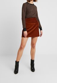 Jupe mini en velours côtelé marron à silhouette ajustée, associée à un haut à manches longues rayé noir et or, et des bottines noires.