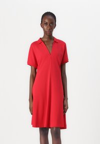 Robe rouge à manches courtes avec un décolleté en V. Fabriquée dans un tissu lisse, elle présente une coupe décontractée et de subtiles fentes latérales pour faciliter les mouvements.