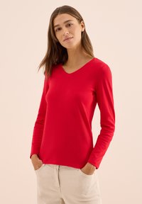 Langärmliges rotes V-Ausschnitt-Shirt aus glattem Stoff, mit einer taillierten Passform und dezenten Nahtdetails. Trägt man zu hellen Hosen.