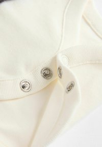 Cremfarbener Playsuit mit glatter Textur, ausgestattet mit metallischen Druckknöpfen und gerippten Kanten am Ausschnitt und den Ärmeln.