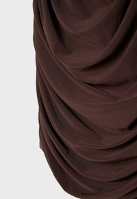 Tumma ruskea kangas, jossa on pehmeät, tasaiset laskokset ja sileä pinta, asetettuna yksinkertaisen vaaleanharmaan taustan eteen.