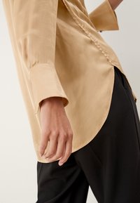 Blouse en satin beige avec un ourlet arrondi et des manches longues, présentant une texture lisse et des détails de couture visibles. Associée à un pantalon noir.