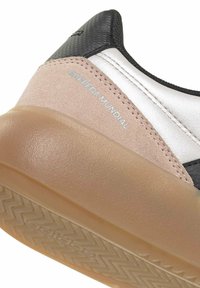 Acercamiento al talón de una zapatilla con ante beige, parte superior negra texturizada, detalle plateado y suela de goma translúcida marcada con "Barreda Mundial".