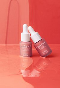 Dos botellas de vidrio de Lumene Liquid Blush con goteros; una es de color rosa claro y la otra de un tono más oscuro, dispuestas frente a un fondo coral reflectante.