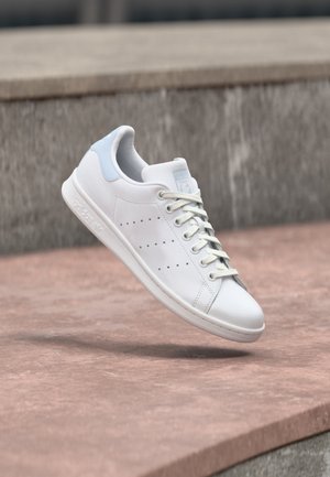 Baskets en cuir blanc avec languette de talon bleu clair, bandes latérales perforées et lacets, flottant au-dessus des marches en pierre en plein air.
