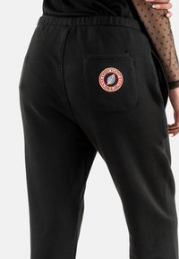 Pantalones de chándal negros con un acabado texturizado, que cuentan con un bolsillo trasero y un logotipo circular con "Sweet Pants" y un diseño de hoja.