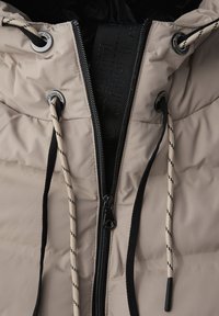Veste matelassée beige avec fermeture éclair noire, cordons noirs et beiges passés à travers des œillets métalliques étiquetés "Street One".