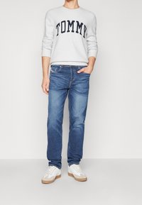 Maglione grigio chiaro con "TOMMY" in blu navy, jeans in denim blu e sneakers bianche con accenti beige. Vestibilità classica, design casual, texture morbida.