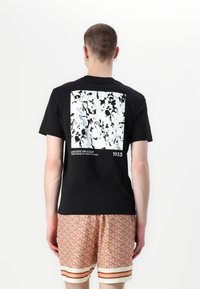 Czarny t-shirt z białą abstrakcyjną grafiką na plecach, z tekstowymi akcentami; noszony z wzorzystymi pomarańczowymi szortami i białym wykończeniem.