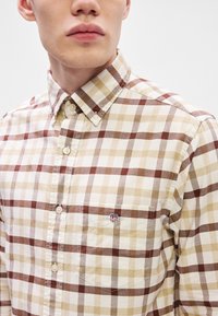 Camicia a quadri beige e marrone con bottoni e taschino sul petto; presenta un piccolo logo ricamato ed è realizzata in tessuto testurizzato.