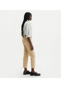 Pantalones de algodón beige con pierna ajustada y dobladillo, combinados con una camisa de botones a rayas blanca y azul marino, usados con mocasines negros.
