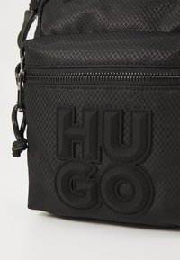 Borsa in nylon nera con superficie texturizzata, dotata di un comparto con zip e un logo "HUGO" in rilievo e audace nella parte anteriore.