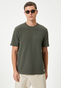 Olivengrøn T-shirt med korte ærmer og struktureret overflade samt rund hals. Bæres med lysebeige bukser og sorte solbriller.