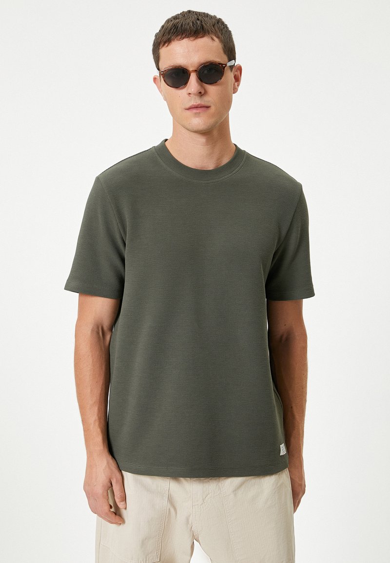Olivengrøn T-shirt med korte ærmer og struktureret overflade samt rund hals. Bæres med lysebeige bukser og sorte solbriller.