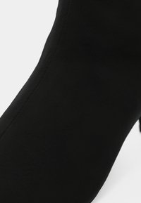 Gros plan d'une botte chevillère en tissu noir montrant la texture sans couture et la forme incurvée sur fond blanc.