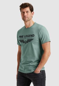 Green cotton t-shirt with a round neck, featuring a black graphic print that includes "PME LEGEND" and wing designs. Dark jeans.