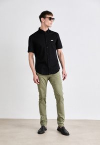 Crna košulja kratkih rukava s gumbima i logom, svijetlozelene uske hlače i crne cipele na slip-on, nosi model protiv jednostavne pozadine.