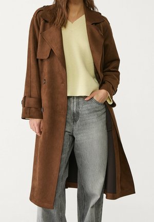 Kobieta nosząca brązowy płaszcz typu trench, jasnożółty sweter z dekoltem w serek oraz wyblakłe szare dżinsy, z jedną ręką w kieszeni.