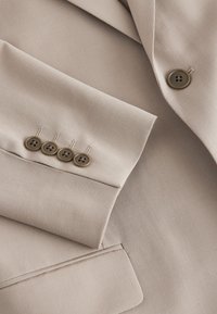 Veste de costume beige avec manchette à quatre boutons, un bouton devant et une poche avant à rabat avec une texture de tissu lisse.