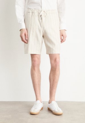 Beige, verticaal gestreepte shorts van lichtgewicht stof, met een aantrekkoord in de taille en zijzakken. Draagt wit sportschoenen.