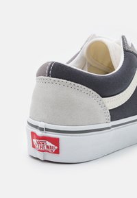 Šedé a bílé semišové tenisky s texturovaným koženým potahem, se výrazným červeným logem Vans na patě a bílou gumovou podrážkou.