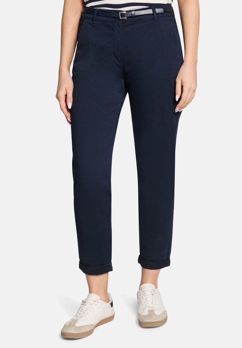 Femme portant un pantalon court bleu marine avec une ceinture noire et des baskets blanches, debout devant un fond uni.