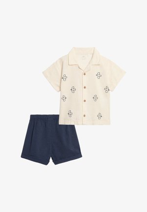 Kurzärmliges cremefarbenes Hemd mit schwarzen Vogelmotiven und Holzknöpfen, kombiniert mit dunkelblauen Shorts mit elastischem Bund.