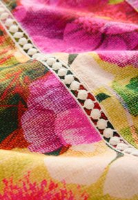 Bunt gemustertes Blumenstoff mit rosa, gelben und grünen Farbtönen und einer strukturierten Oberfläche. Enthält einen weißen Pomponbesatz entlang der Naht.