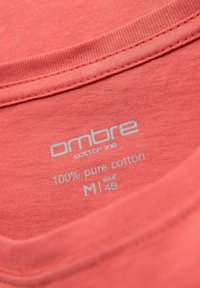 Štítek na korálovém bavlněném tričku s světle šedým textem. Zní: "ombre bavlněná řada 100% čistá bavlna M eur 48." Hladká textura, kulatý výstřih.