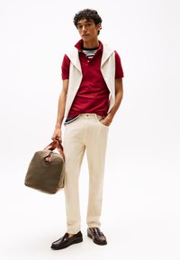 Rdeča polo majica pod krem puloverjem brez rokavov, krem hlače, rjavi loaferji, v roki drži kaki torbo z rjavimi trakovi.
